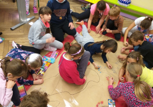 Uczniowie siedzą na podłodze i tworzą wspólną prace na kartonie papieru.